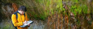 A man in a yellow jacket records information in a notebook, standing near a moss covered wall